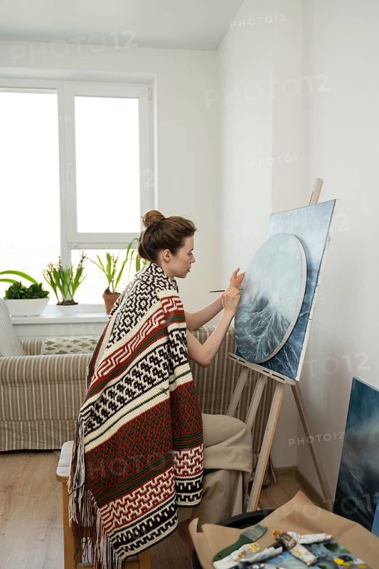 An artist, wrapped in a colorful blanket, paints on a circular canvas in a bright home studio, creating a tranquil and inspiring atmosphere for creativity