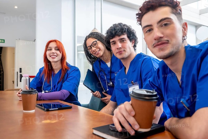 Diverse group of young medical students in blue scrubs with stethoscopes, sharing a coffee break and smiling at camera in a hospital or campus setting, relaxed and friendly colleagues