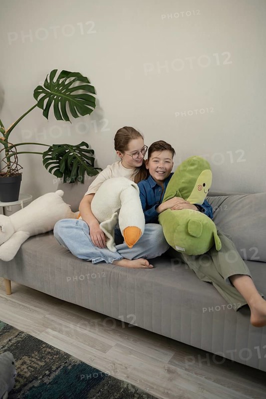A joyful pair of siblings sitting on a plush sofa with stuffed toys, sharing smiles and laughter. The room features a modern potted plant and stylish decor