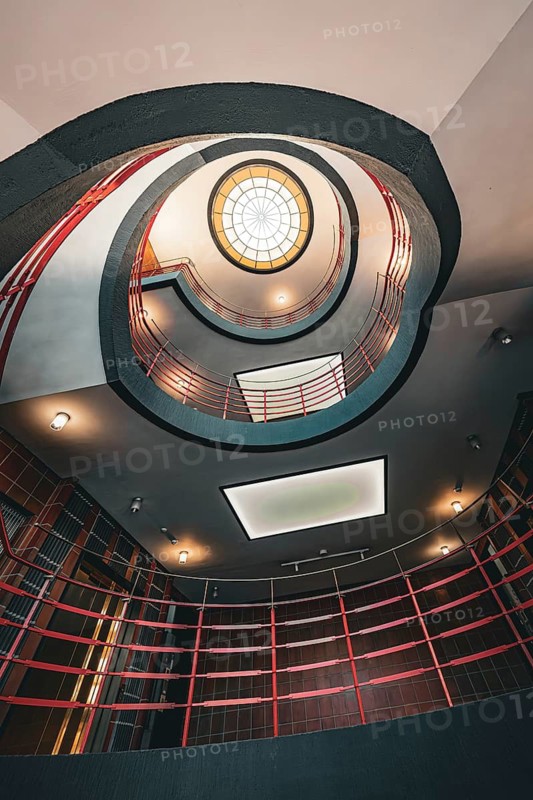 View upwards through a spiral staircase with red banisters and round ceiling lights, SPRINKENHOF Hamburg, Germany