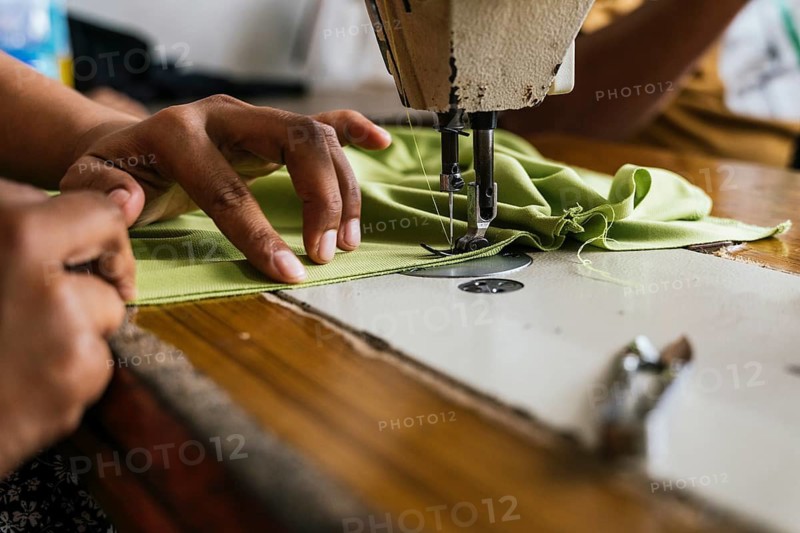 A close up shot of a sewing machine with asian seamstress hands skillfully guiding green fabric through the needle highlights craftsmanship and textile work in action