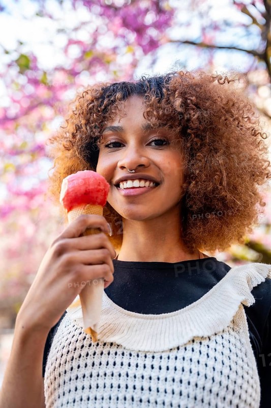 Smiling young woman enjoying an ice cream cone while standing under a blooming tree, surrounded by vibrant flowers on a sunny spring day