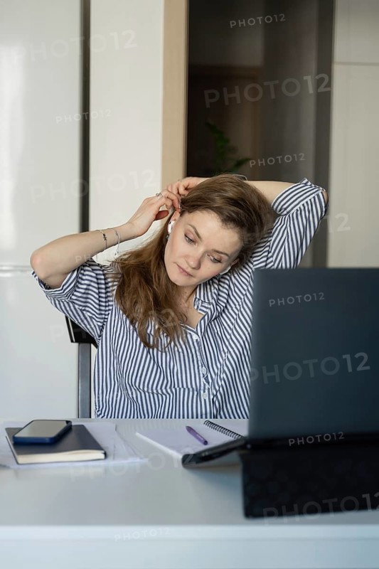 Woman in a striped shirt at home, adjusting her earphones while working on a laptop. A phone, notebook, and pen are on the table, creating a cozy workspace