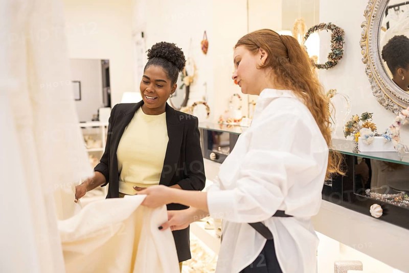 Two women are joyfully examining a wedding dress in an elegant boutique. The bright interior enhances the atmosphere of excitement and decision making