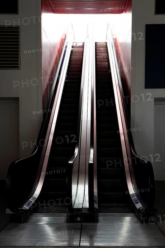 A starkly lit escalator captured between moments of use, creating a sense of movement amidst a sleek, modern urban architecture