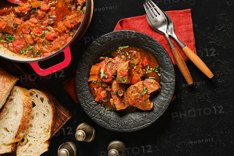 A bowl of beef stew sits on a black surface next to crusty bread. The stew has meat, vegetables, and herbs. A cooking pot is nearby, along with forks and a red napkin