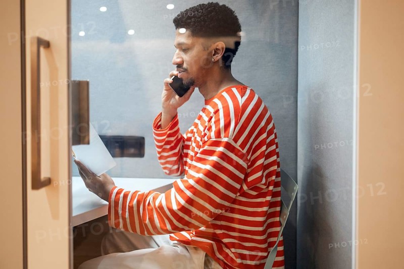 A professional seated in a private coworking booth making a phone call while holding a document. The setting is modern, with soft lighting and a soundproof environment