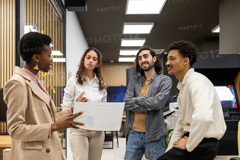 A diverse group of professionals engaged in a team meeting in a modern office. They are discussing a project, with one person holding a document, indicating active collaboration