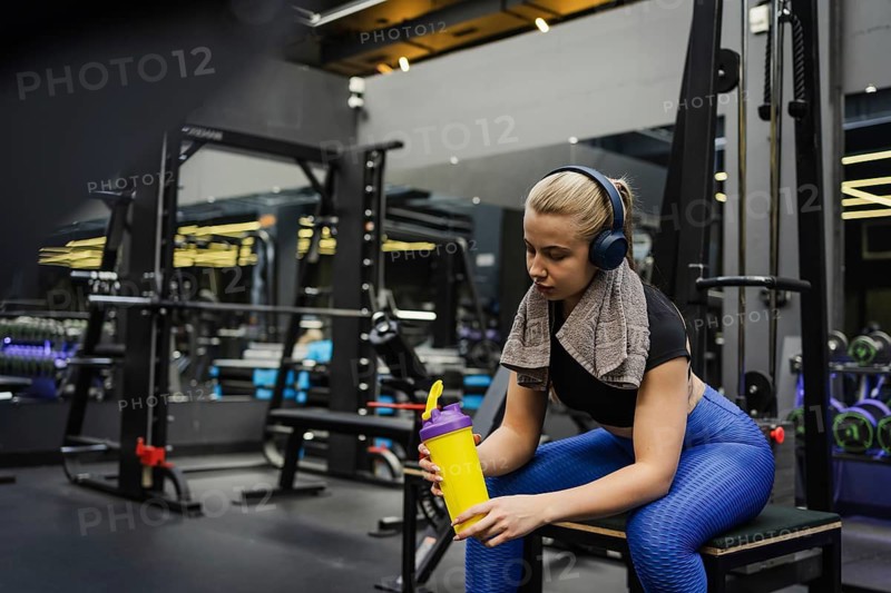 A blonde woman in athletic wear relaxes on a bench in a modern gym, holding a yellow protein shaker. She wears headphones and a towel around her neck, enjoying a break from exercise