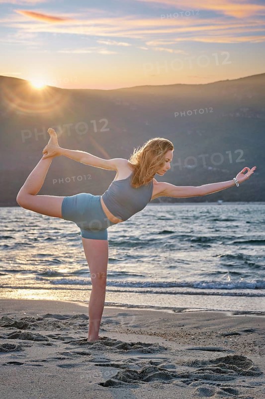 A woman performs Natarajasana, or Dancer's Pose, on a sandy beach at sunset. She balances gracefully as the sun sets behind the mountains, creating a serene atmosphere