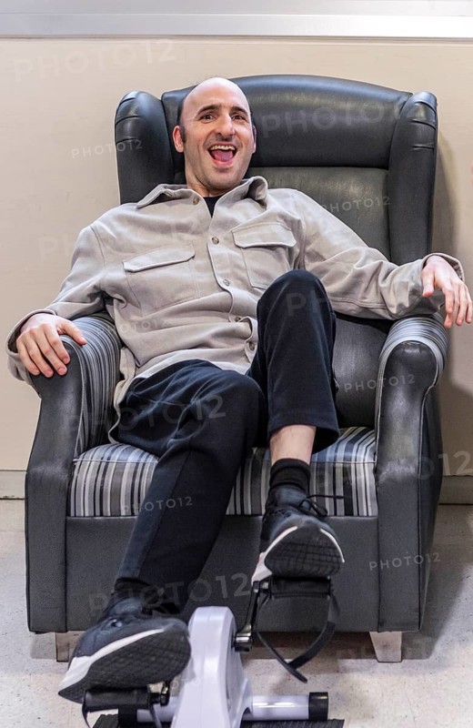 A man enjoys relaxation and exercise on a chair with a pedal machine in a mental health residence. The scene captures a balance of leisure and physical activity in a supportive environment