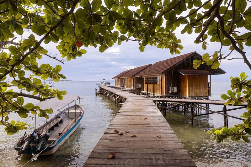 A serene view of wooden huts on stilts and a moored boat framed by lush leaves on Waigeo Island, part of the Raja Ampat Archipelago, West Papua, Indonesia