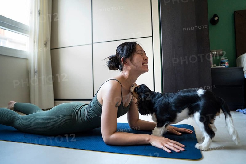 Young Asian woman attempting to exercise at home on a yoga mat while her playful dog interrupts her routine, bringing a joyful and amusing moment to the workout session