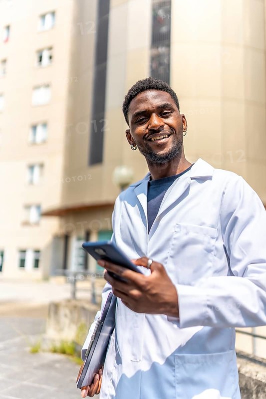 Vertical photo of a cheerful young african doctor using phone standing outside the hospital