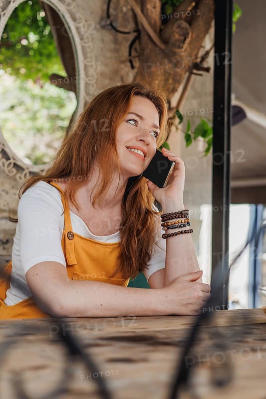 A cheerful woman tourist in her 40s, wearing an orange dress, enjoys a sunny day in an old city in Cesme, Turkey, using her smartphone during her vacation