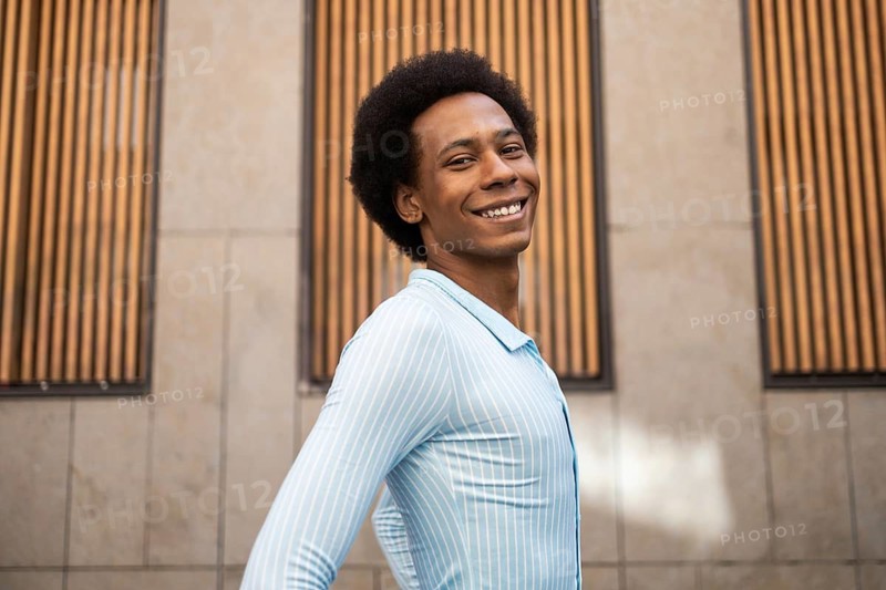 A person with a bright smile poses confidently in front of a contemporary building with wooden panels, radiating positivity and style in a casual blue shirt