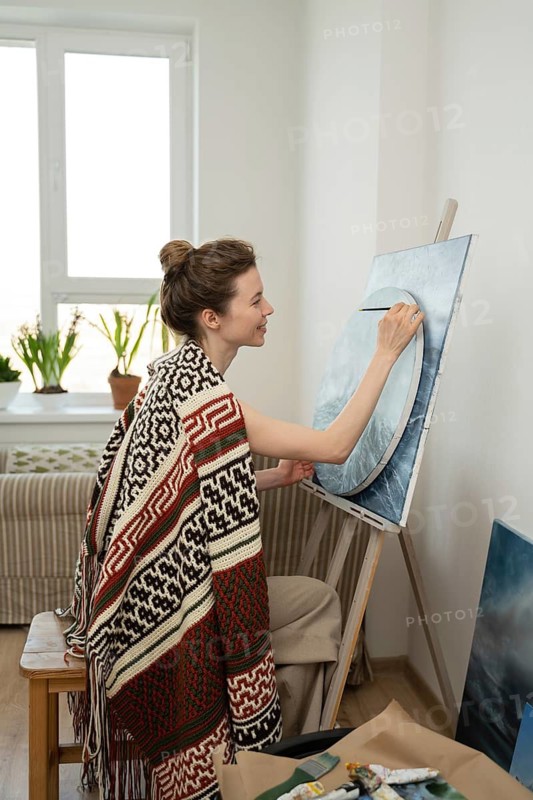 A woman artist paints on a canvas in a cozy room with natural light streaming through a window, creating a serene and focused atmosphere