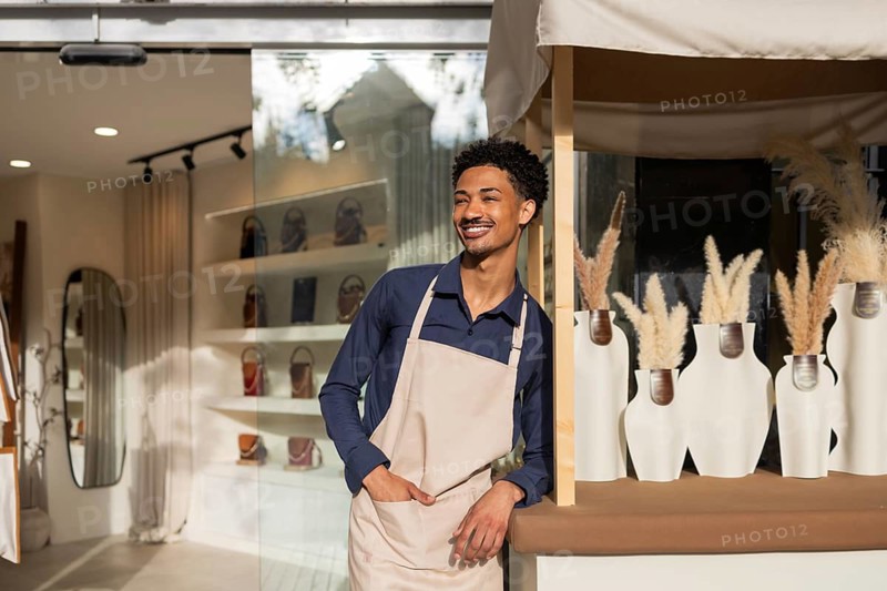A cheerful shopkeeper in an apron leans against a counter in a trendy bag store. The stylish interior features a display of bags and elegant decor under soft lighting