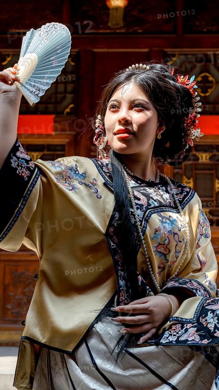A young woman in Qing Dynasty attire poses elegantly in Pingyao, China, holding a delicate fan. Her traditional clothing is richly decorated, embodying historical Chinese fashion