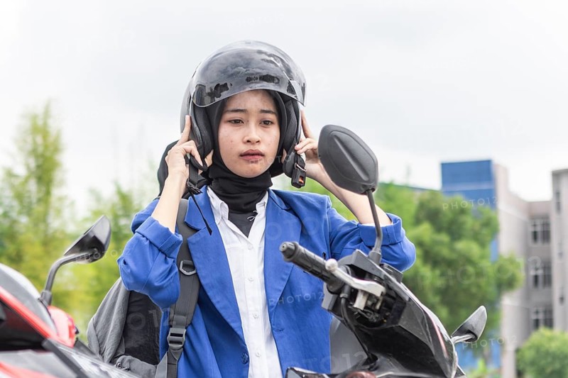 A young Asian Muslim woman wearing a blue jacket and helmet stands by her scooter, getting ready for a commute. The image depicts the theme of returning to school