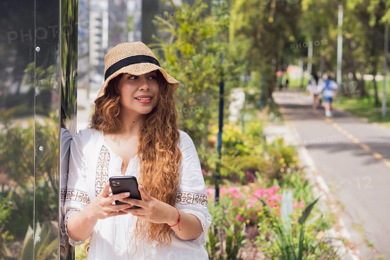 A young American Latin woman enjoys a sunny day outside, leaning against a reflective surface and holding a smartphone, surrounded by lush greenery and vibrant flowers