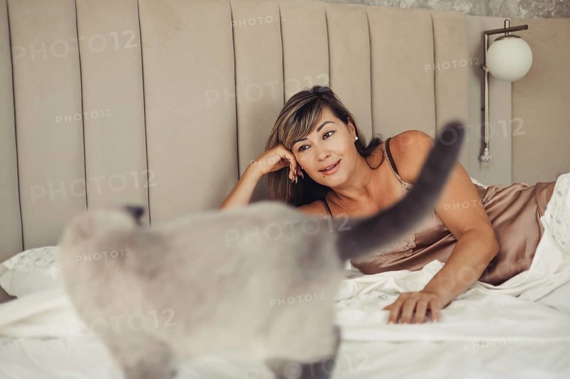 A woman in a satin nightgown lies on a bed, smiling contentedly at her pet cat. The cozy room features soft lighting and a plush headboard, creating a relaxed atmosphere