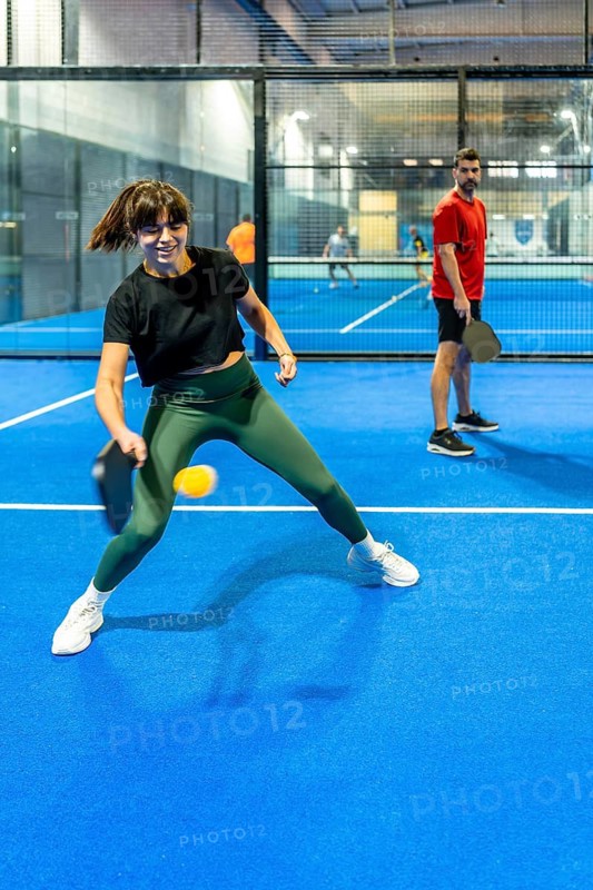 Woman striking a pickleball with a paddle, actively engaged in a fast paced game on an indoor court, with another player standing in the background, focusing on leisure and fitness