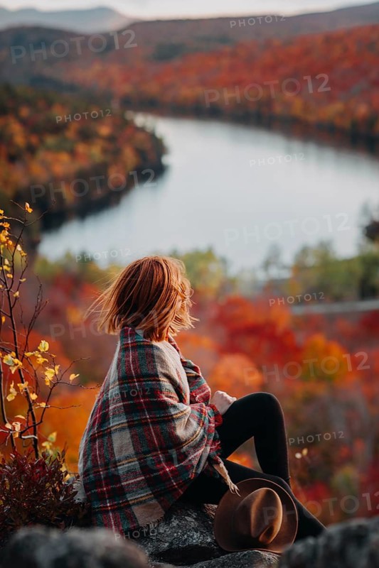 A woman draped in a cozy plaid shawl sits on a rock overlooking a stunning autumn landscape in Quebec, Canada. The vibrant fall foliage complements the serene setting