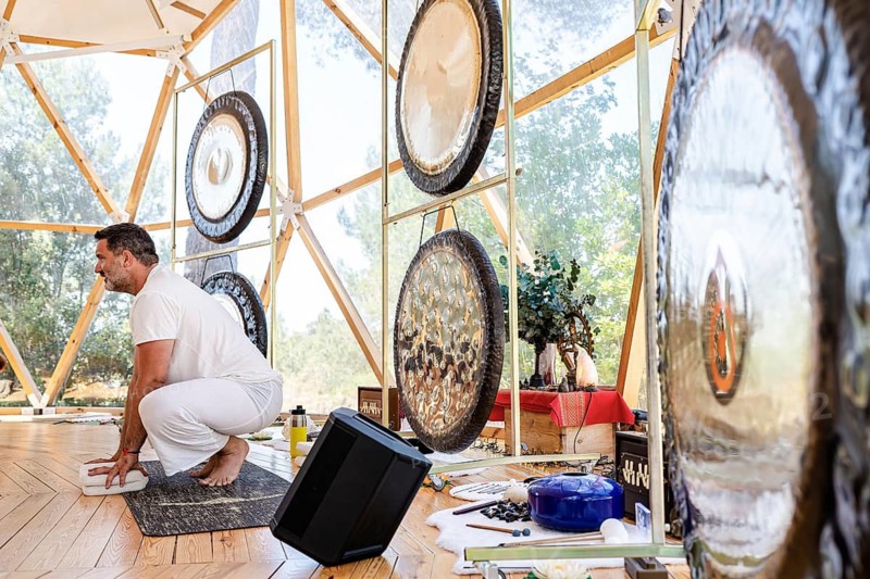 A male sound therapist in a serene geodesic dome setting prepares for a sound therapy session with various gongs and sound tools, creating a calming atmosphere for relaxation
