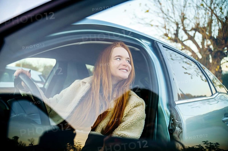 A beautiful pregnant woman with red hair sits comfortably in a car, looking outside with a peaceful expression. The natural light and outdoor setting create a serene atmosphere