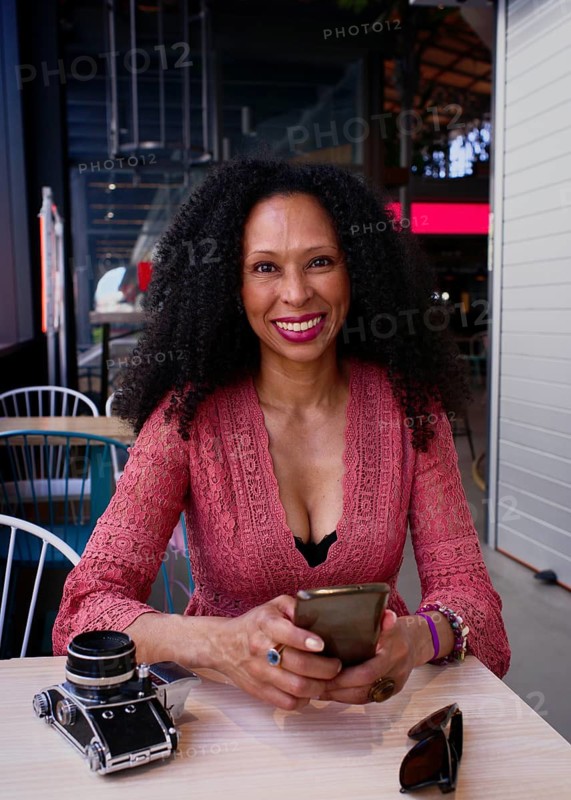 A latin woman with curly hair sits at cafe, smiling warmly at the camera. She holds a smartphone, with a vintage camera and sunglasses on the table