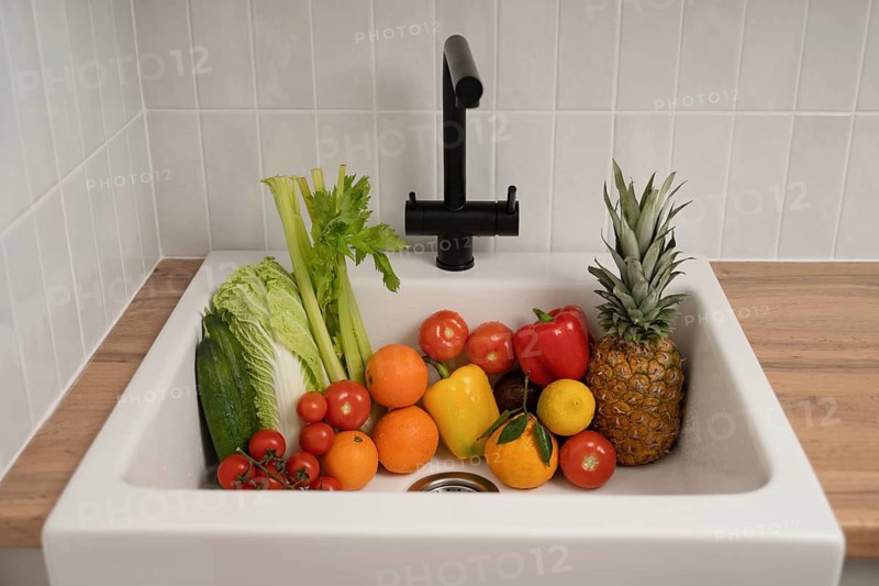 A vibrant collection of fresh fruits and vegetables, including tomatoes, celery, bell peppers, and pineapple, arranged in a kitchen sink with a modern black faucet