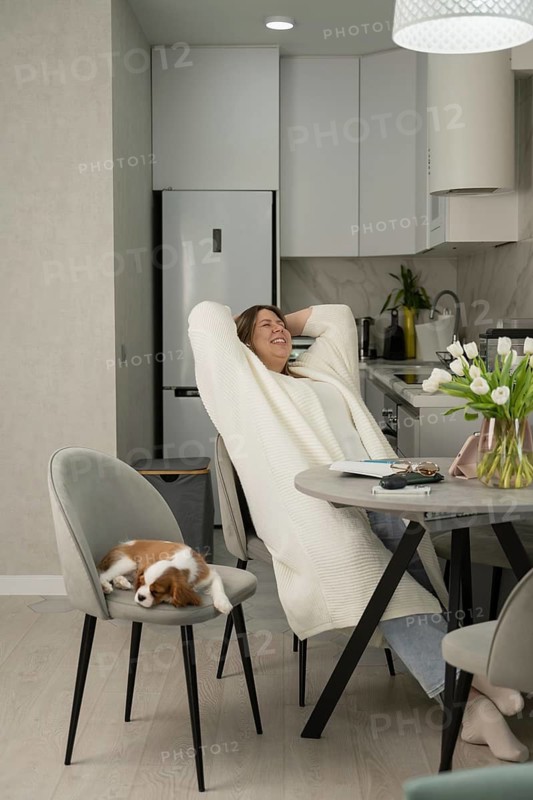 A woman relaxes in a modern kitchen, smiling while sitting at a table. A small dog sleeps on a chair beside her. A bright and tranquil home setting