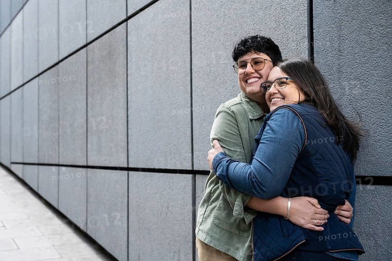 A joyful lesbian couple shares a warm embrace outdoors, standing against a modern urban wall. The happiness is evident on their smiling faces as they enjoy their moment