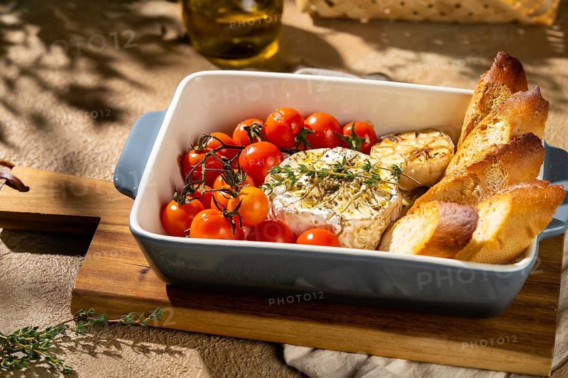 A baked Camembert cheese dish, paired with roasted cherry tomatoes, garlic, and crispy baguette slices in a ceramic dish. Enjoy the flavors and textures of this gourmet delight