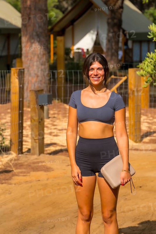 Young woman smiling and holding her purse while standing on a dirt path at a glamping resort, surrounded by canvas tents and enjoying the sunny outdoor atmosphere