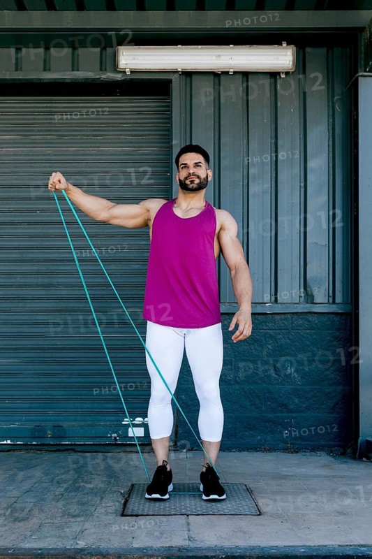Fit man performs outdoor resistance band workout. Wearing vibrant tank top and leggings, he demonstrates strength and fitness on a sunny day, focusing on arm muscles