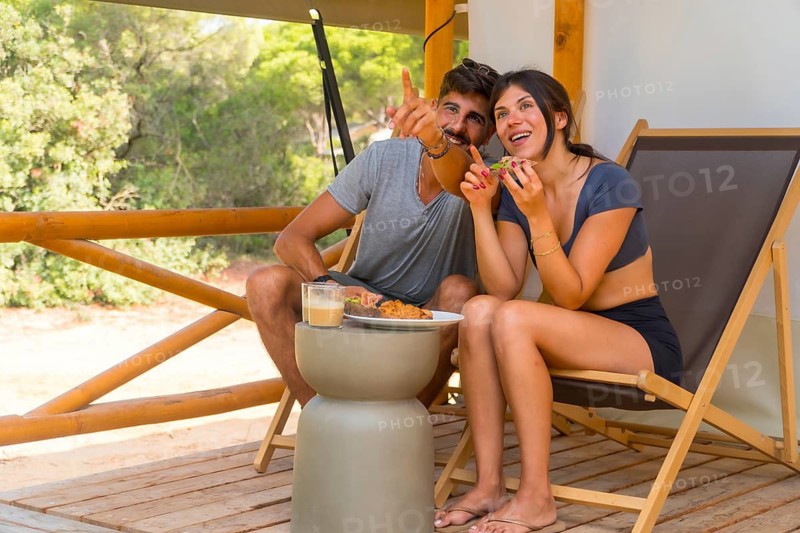 Happy couple eating breakfast and pointing at something while sitting on the wooden patio of their glamping tent, enjoying the morning sunlight and nature