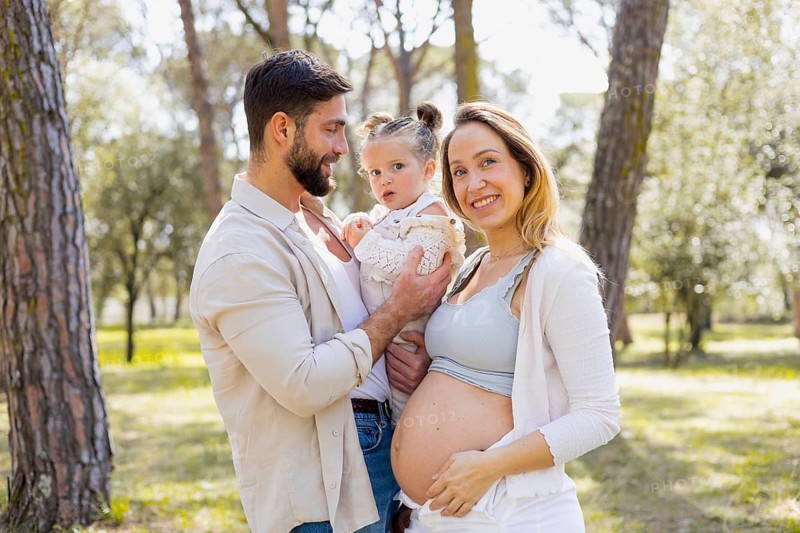 A pregnant woman joyfully stands with her family in a sunny outdoor setting, capturing a moment of happiness and connection with her partner and young child in nature