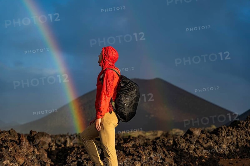 An adventurer in a red jacket and backpack explores the volcanic terrain of Timanfaya National Park, Lanzarote. A vibrant rainbow arcs across the dramatic sky