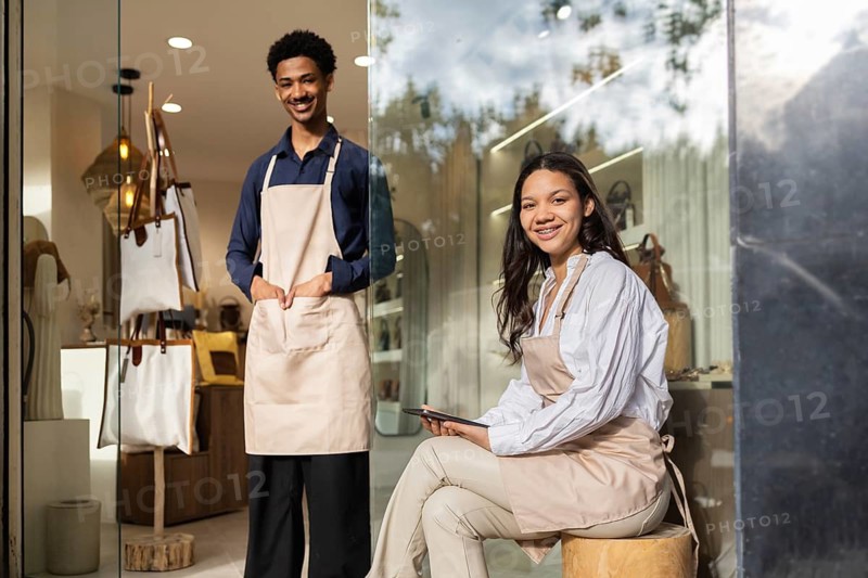 Two smiling shop assistants in a chic bag store, both wearing aprons. One standing, the other seated, ready to assist customers with their shopping experience