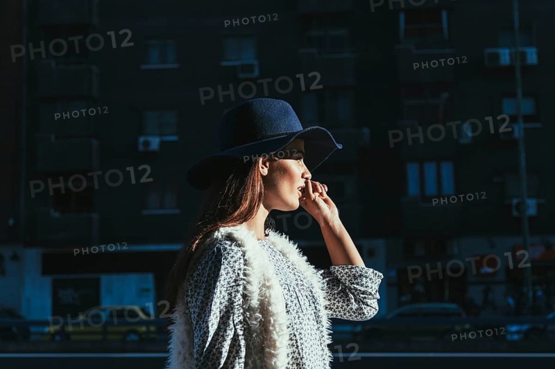 A woman in a chic hat and stylish attire enjoys a sunny morning in the city. The urban landscape and her thoughtful expression add a sense of sophistication and tranquility