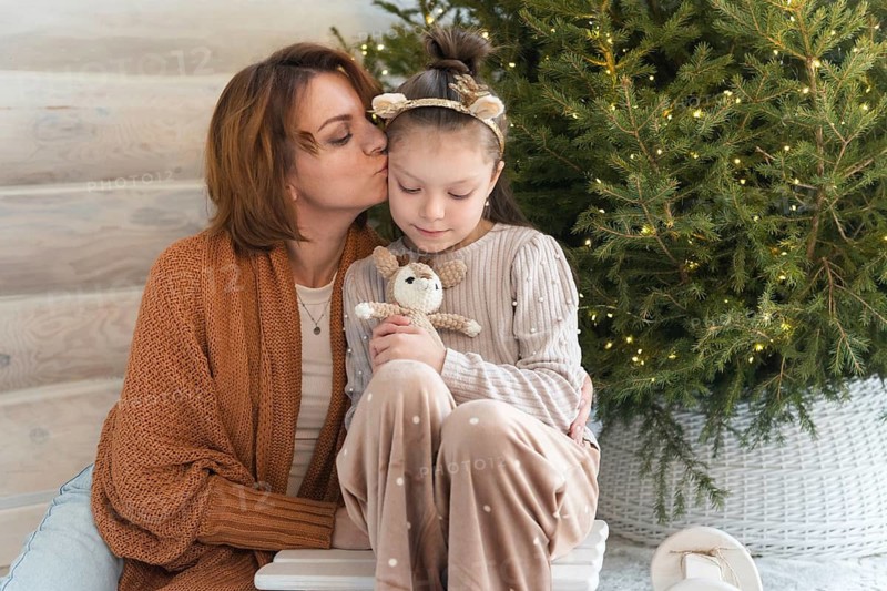 A mother lovingly kisses her daughter beside a festively decorated Christmas tree. The young girl, holding a plush toy, is looking down while her mother gazes at her with affection, capturing the spirit of family and holiday celebrations