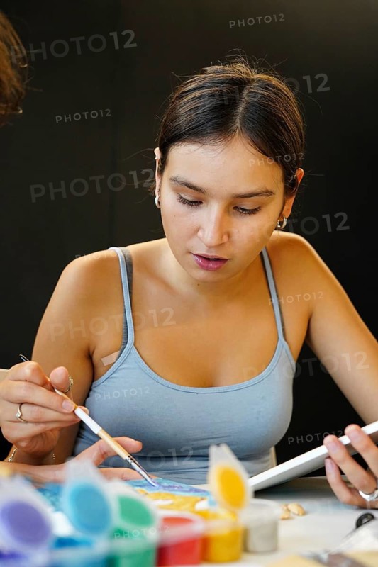 A teenage girl is intently engaged in a painting activity in an art class, using a brush, with a concentration that reflects her artistic involvement. This moment captures her creativity and educational engagement in an art class setting