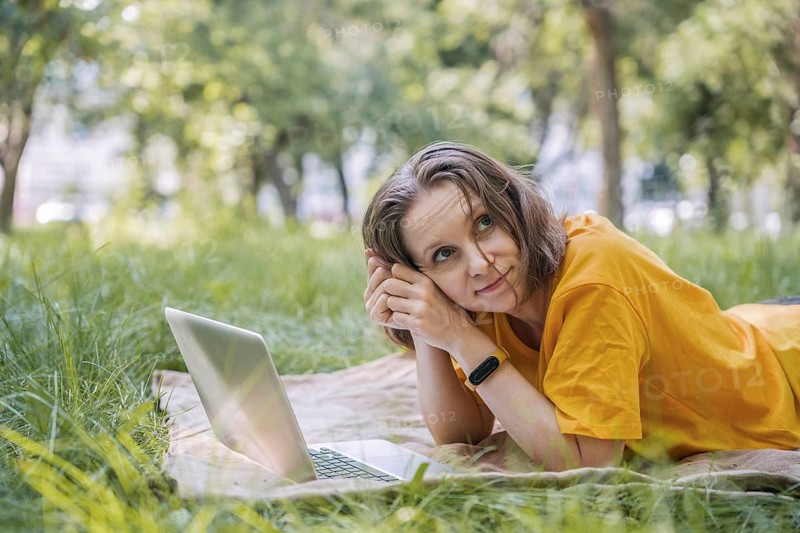 A woman lies comfortably on a blanket in a sunny park, using her laptop. She enjoys the tranquility of nature while engaging in remote work or leisure activities