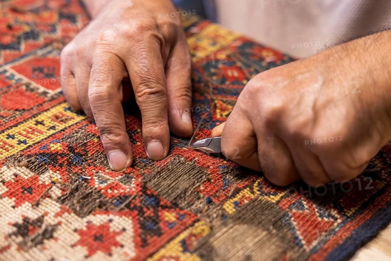 Cropped unrecognizable senior master craftsman from Iran meticulously restores a vibrant, traditional carpet in his workshop located in Salzburg, Austria. His skilled hands wielding a small tool carefully over the fabric's intricate patterns