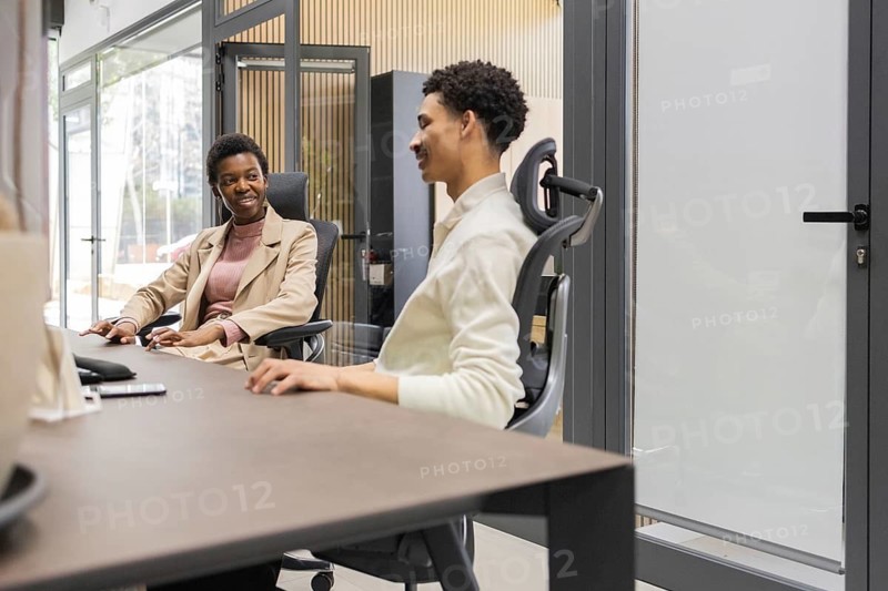 Two colleagues converse in a modern office space, seated comfortably in ergonomic chairs. The environment is bright, fostering a collaborative and professional atmosphere