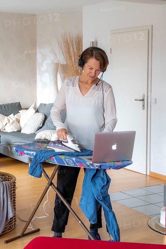 Woman, mid-50s, works from home, with laptop and communicates with colleagues via headset, home office, irons laundry in parallel