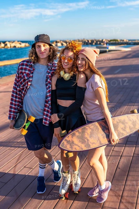 Three happy skater friends posing together on a boardwalk by the sea, holding skateboards and roller skates