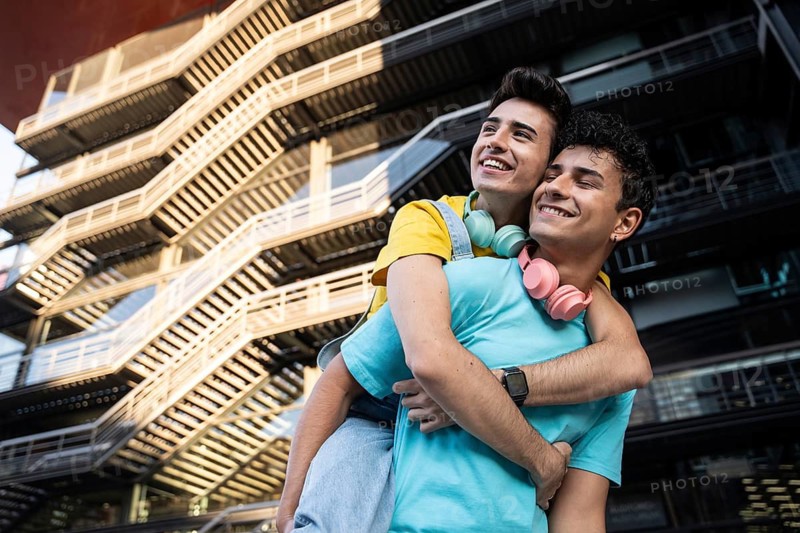 A happy gay couple shares a joyful piggyback ride against a modern urban backdrop. Their genuine laughter and casual attire embody love and happiness in the city